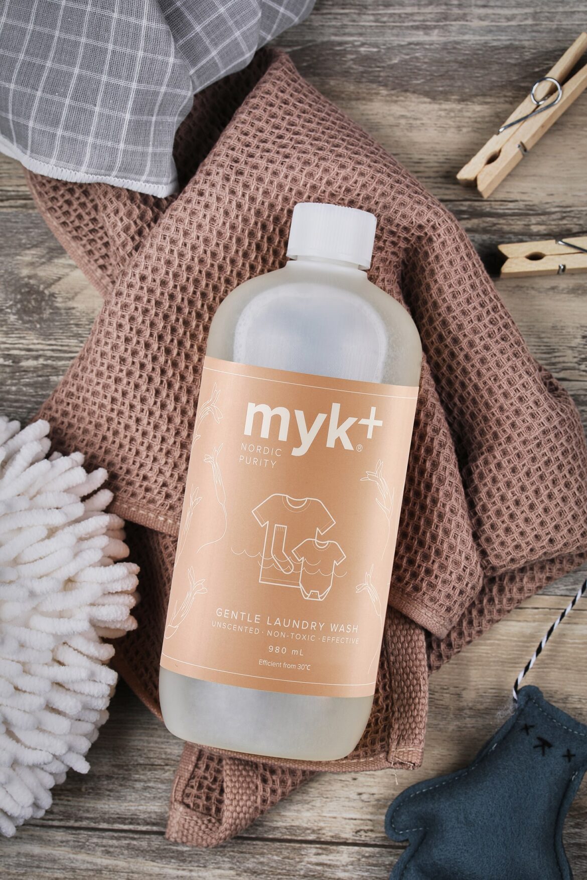 Бутылка средства для бережной стирки myk+ на коричневой фактурной ткани. На этикетке изображена одежда на веревке. Рядом на деревянной поверхности — белая скребница, серая ткань, прищепки и перчатка.
