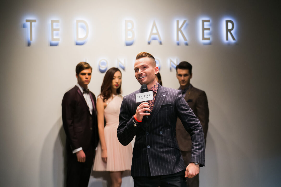 Мужчина в полосатом костюме говорит в микрофон перед вывеской Ted Baker London. Три модели, двое мужчин и одна женщина, стоят позади него в официальных нарядах.