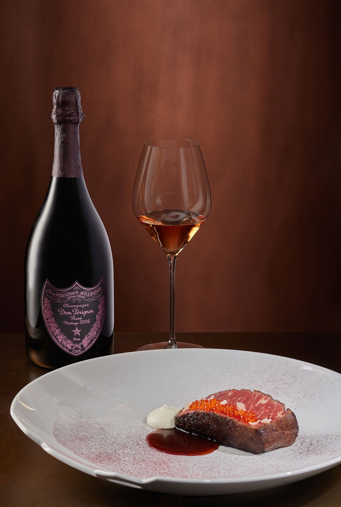 Бутылка вина Dom Pérignon стоит рядом с наполненным бокалом вина. На переднем плане — изысканное блюдо с ломтиком мяса, увенчанным оранжевой икрой, ложкой сливок и соусом, все на белой тарелке на коричневом фоне.
