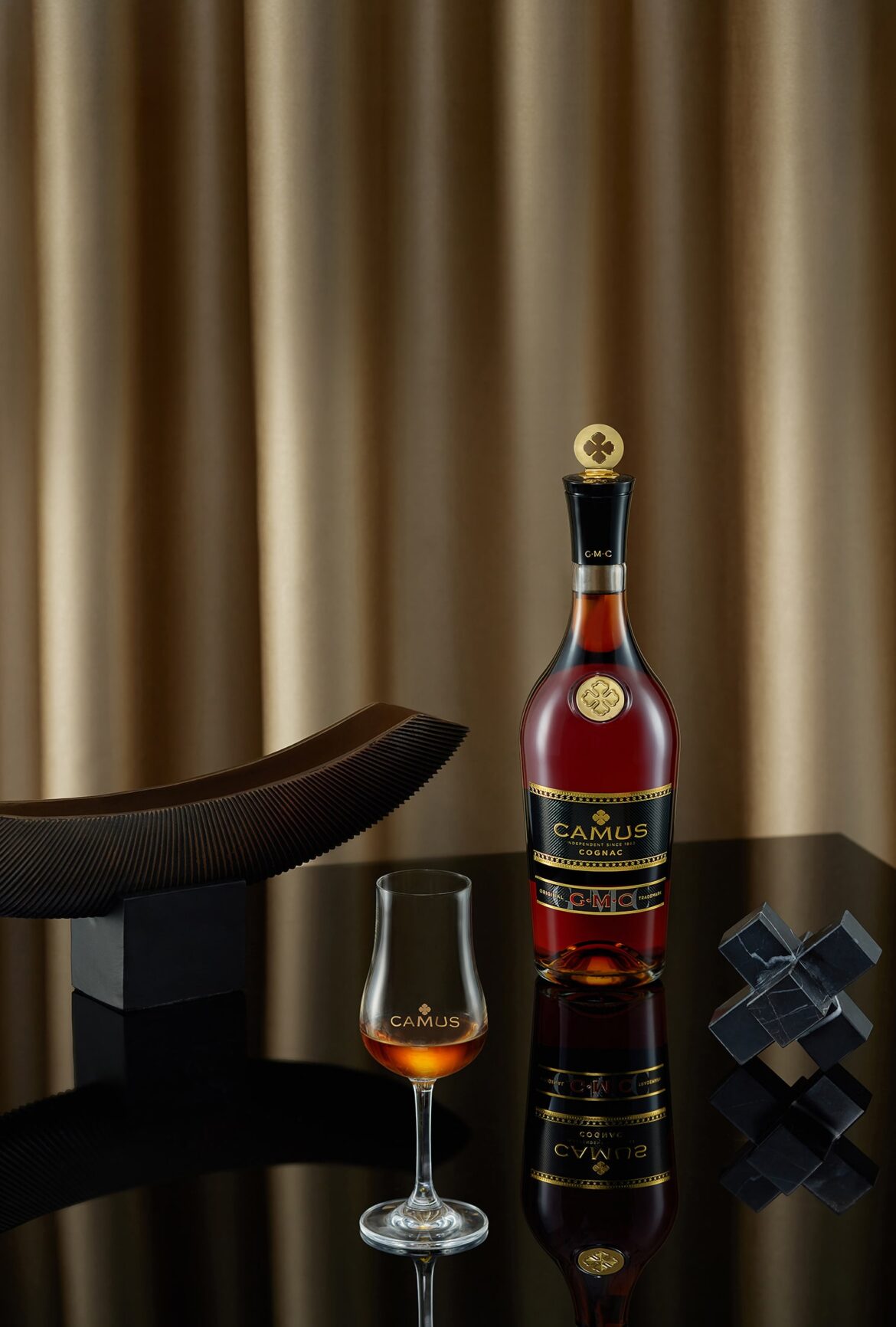 Бутылка коньяка Camus стоит на отражающей черной поверхности, рядом с наполненным бокалом. Рядом находятся две абстрактные черные скульптуры. На заднем плане — золотисто-драпированные занавески.