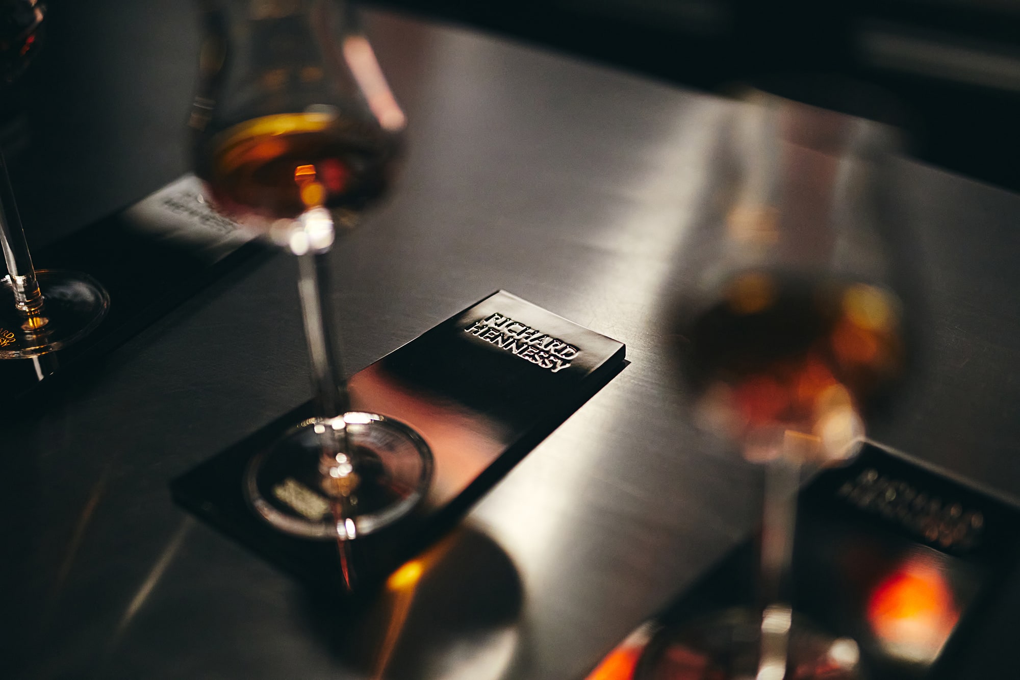 Тускло освещенный стол с элегантными бокалами и карточкой с текстом Richard Hennessy. Сцена передает изысканную атмосферу.