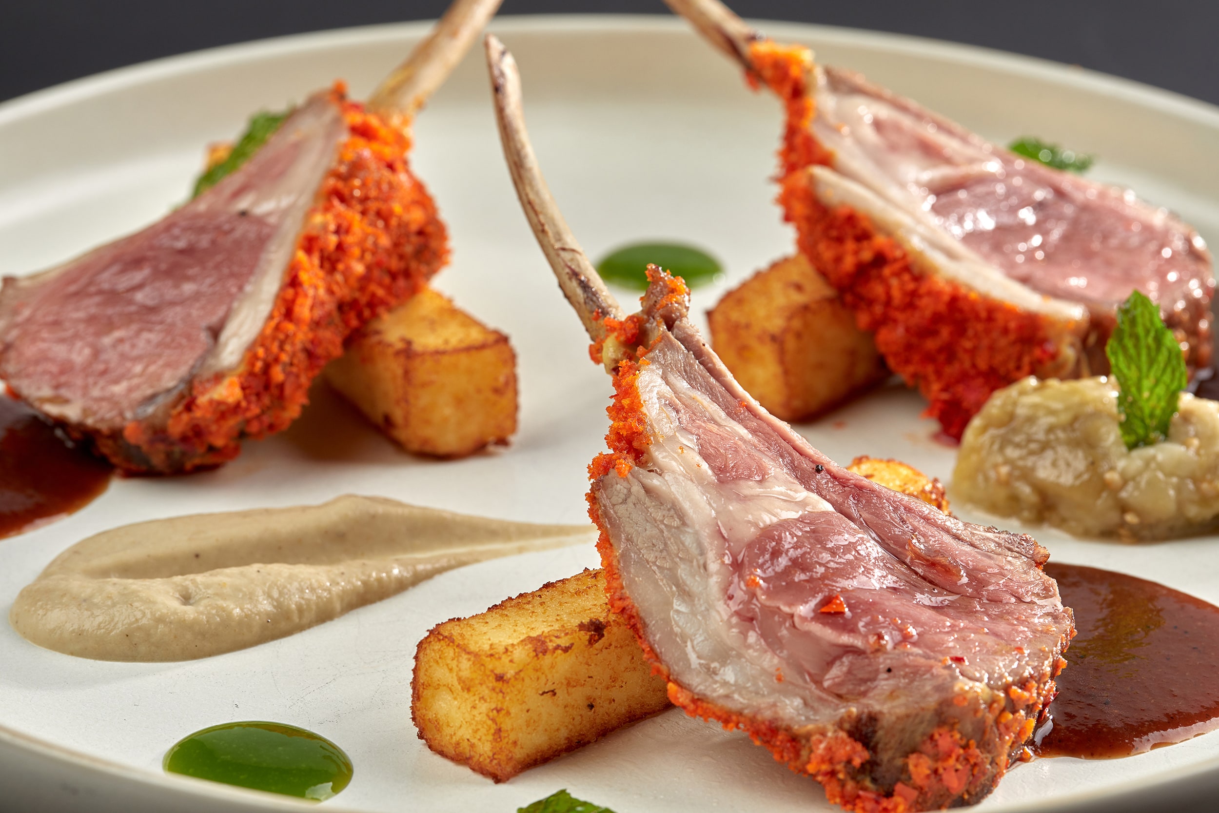 Каре ягнёнка в панировке с картофелем и соусом на белой тарелке