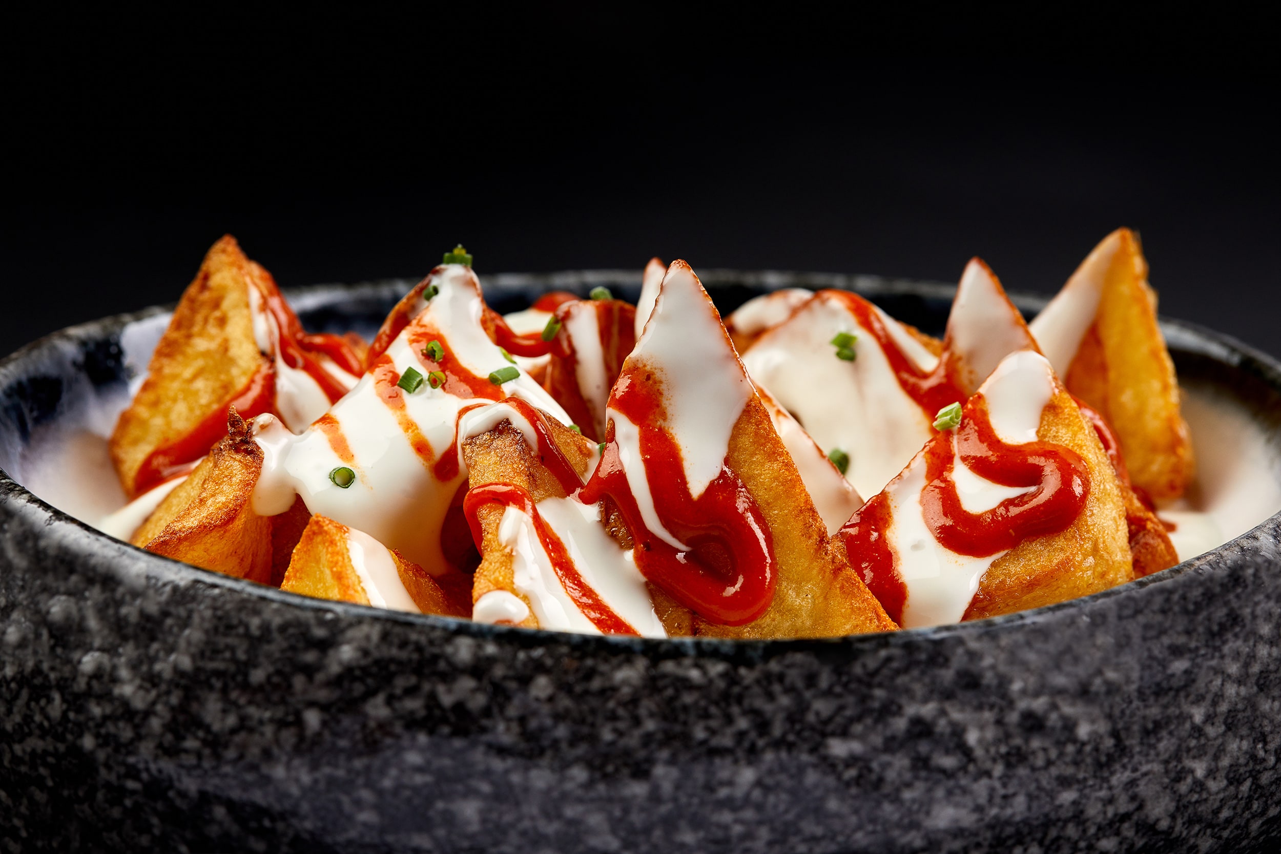 Запечённый картофель с белым и красным соусом в каменной миске на чёрном фоне