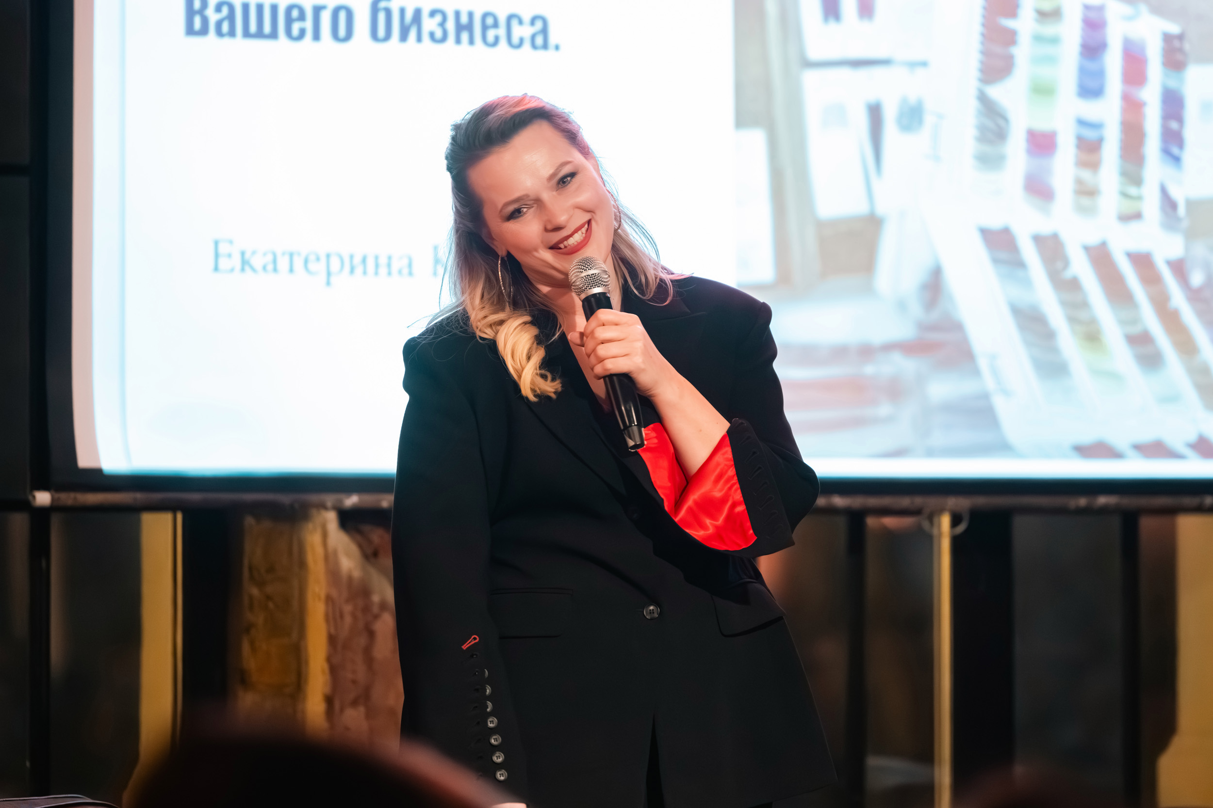 Женщина в черном пиджаке стоит на сцене, держа в руках микрофон, и улыбается, когда говорит. За ее спиной - экран, на котором отображается текст на русском языке и разноцветные предметы, что наводит на мысль о презентации или лекции. Фото из портфолио фотографа Павла Шубского.