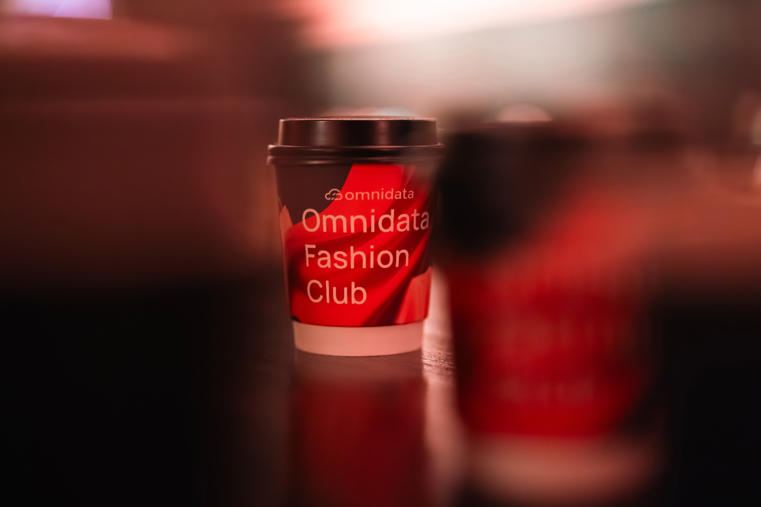 Стаканчик с кофе на вынос с красно-белой надписью Omnidata Fashion Club стоит на отражающей поверхности, на переднем плане - еще один размытый стаканчик. Фон мягко освещен и не сфокусирован. Фото из портфолио фотографа Павла Шубского.
