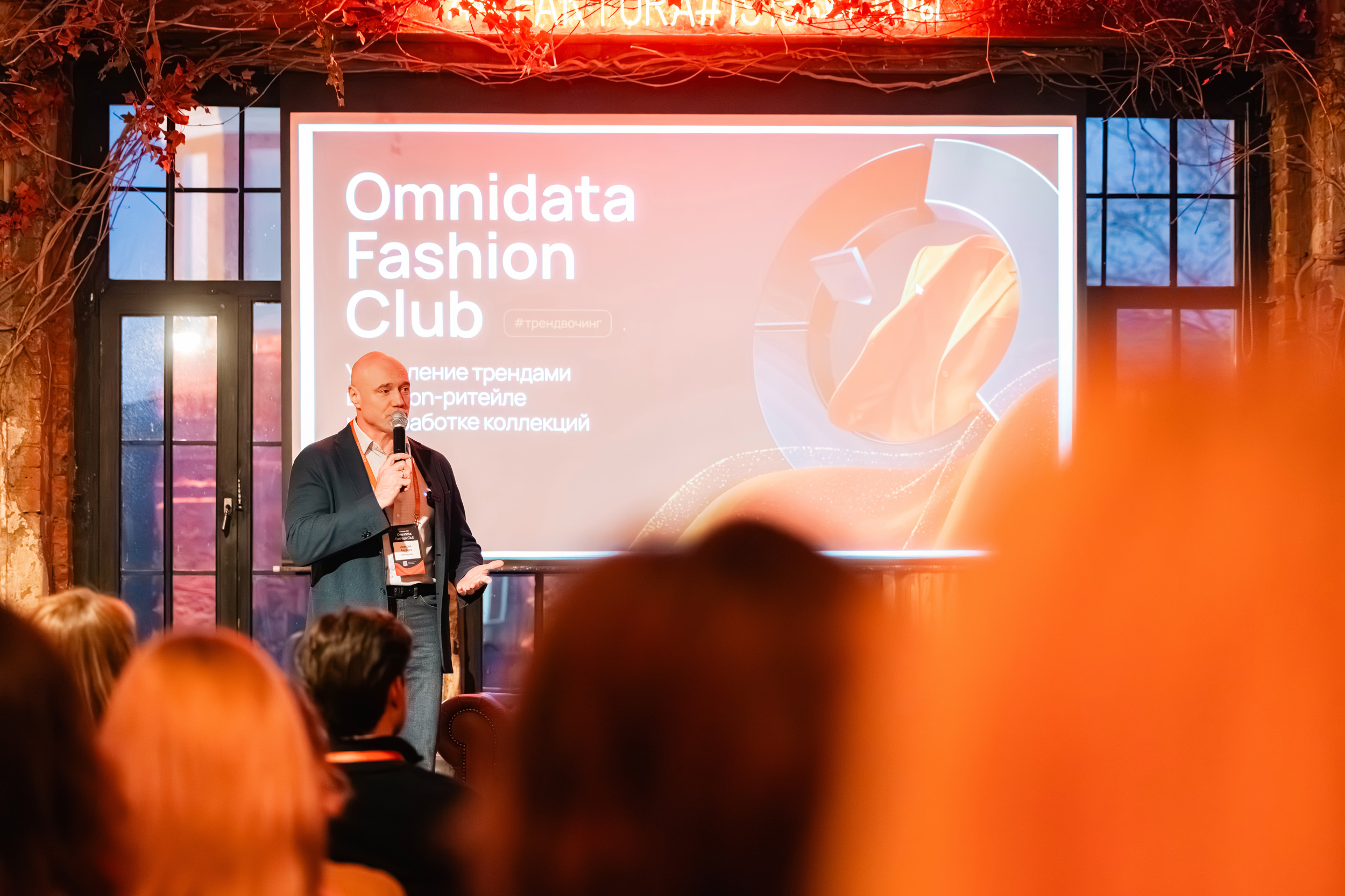 Мужчина с микрофоном выступает на сцене перед экраном с изображением Omnidata Fashion Club на закрытом мероприятии, а зрители слушают. Фото из портфолио фотографа Павла Шубского.