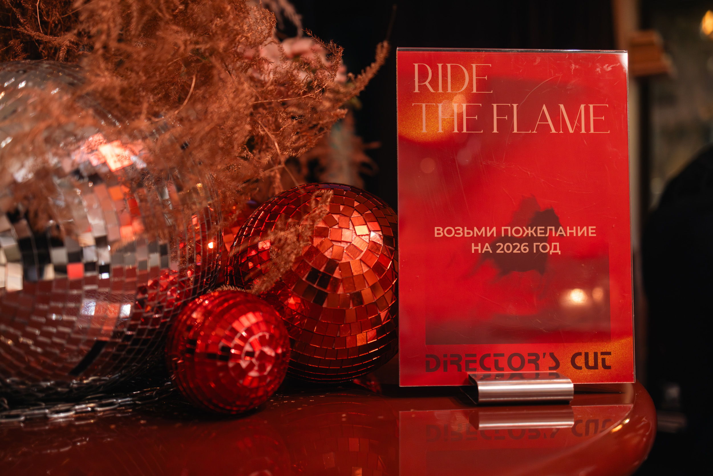 Блестящий диско-шар и красные орнаменты рядом с красной табличкой с надписью RIDE THE FLAME и ВОЗЬМИ ПОЖЕЛАНИЕ НА 2026 ГОД с надписью DIRECTORS CUT внизу, все на отражающей поверхности. Фото из портфолио фотографа Павла Шубского.