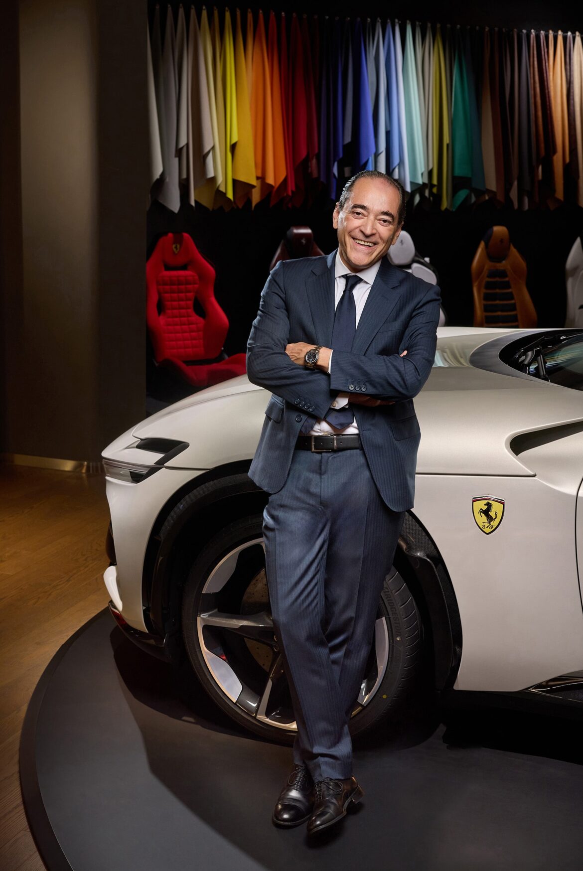 Мужчина в костюме прислонился к белому роскошному автомобилю с логотипом Ferrari. За ним выставлены образцы тканей разных цветов. Он улыбается, скрестив руки, стоя в выставочном зале с деревянным полом. Мужчина в костюме прислонился к белому роскошному автомобилю с логотипом Ferrari. За ним выставлены образцы тканей разных цветов. Он улыбается, скрестив руки, стоя в выставочном зале с деревянным полом.