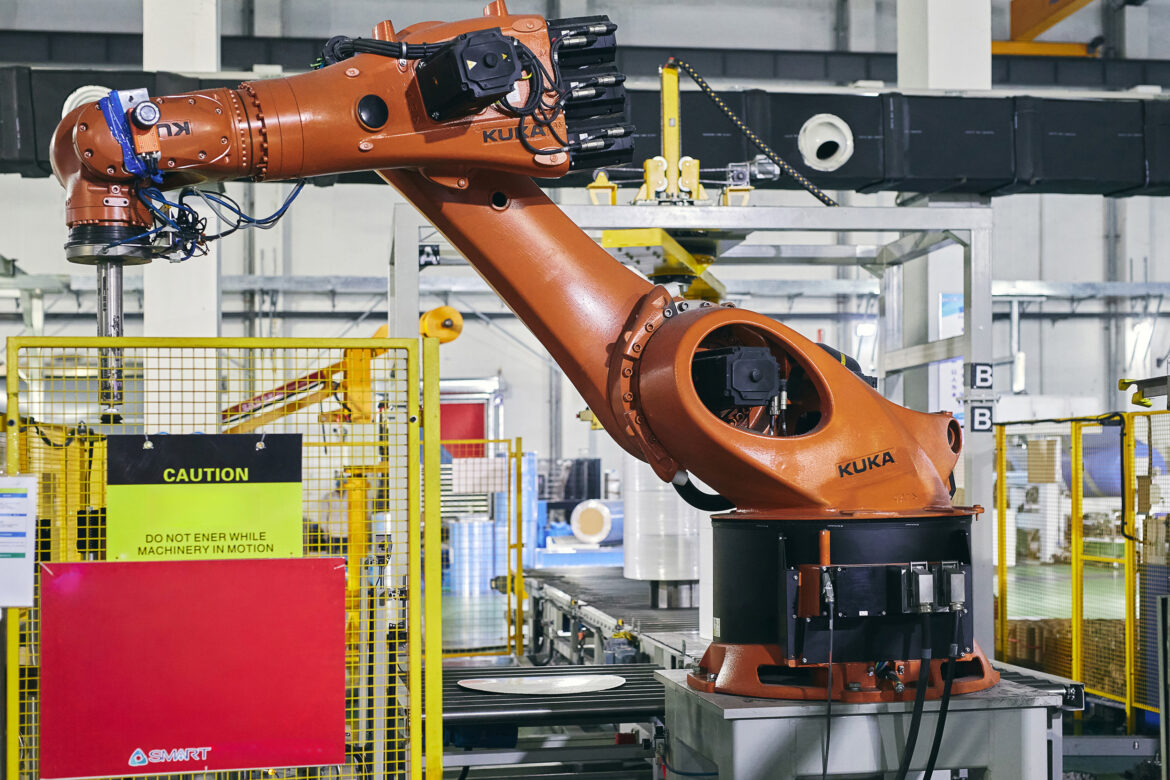 Промышленный оранжевый роботизированный манипулятор с надписью KUKA работает на заводе вблизи защитных ограждений и знака, предупреждающего о движущемся оборудовании. На заднем плане видны металлические конструкции и оборудование. Фото из портфолио фотографа Павла Шубского. Промышленный оранжевый роботизированный манипулятор с надписью KUKA работает на заводе вблизи защитных ограждений и знака, предупреждающего о движущемся оборудовании. На заднем плане видны металлические конструкции и оборудование. Фото из портфолио фотографа Павла Шубского.