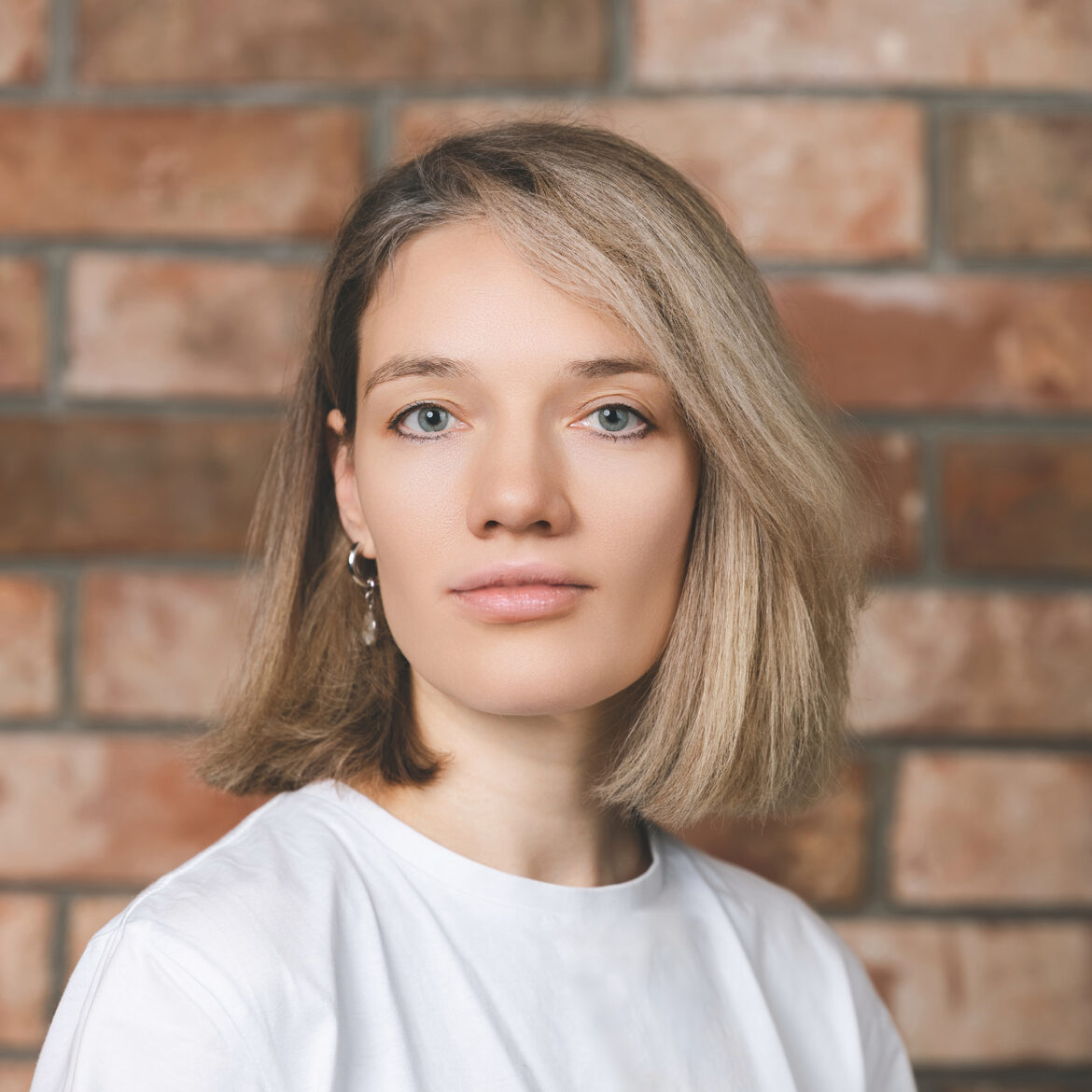 Молодая женщина с прямыми светлыми волосами до плеч и голубыми глазами, одетая в белый топ и серьги-подвески, стоит перед кирпичной стеной и спокойно смотрит в камеру. Фото из портфолио фотографа Павла Шубского. Деловой портрет девушки на фоне кирпичной стены — корпоративная фотосессия в офисе Фото из портфолио фотографа Павла Шубского.